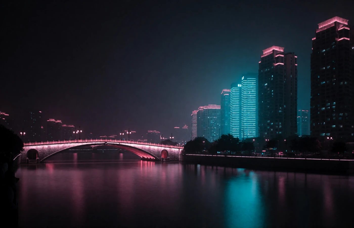 Night view of a Chinese metropolitan waterfront with an illuminated stone arch bridge and modern towers reflected in the river